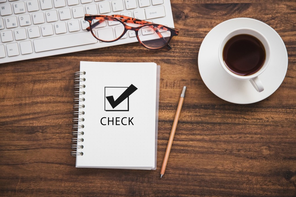 木目のデスク上に置かれた「CHECK」の文字とチェックマークが印刷されたノート、鉛筆、メガネ、キーボード、コーヒーカップ