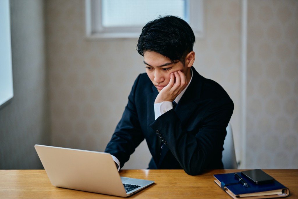 ノートパソコンの前で頬杖をつき悩むスーツの男性