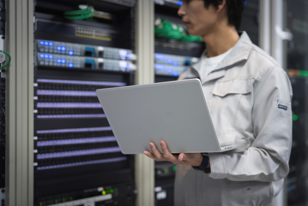 サーバーラックの前でノートパソコンを持つ作業着のエンジニア