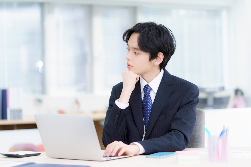 ノートPCの前で顎に手を当てて考え込むスーツ姿の男性社員